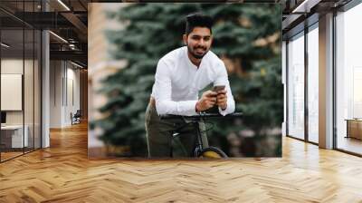 Chilling after good ride. Handsome young indian man holding mobile phone while standing near his bicycle Wall mural